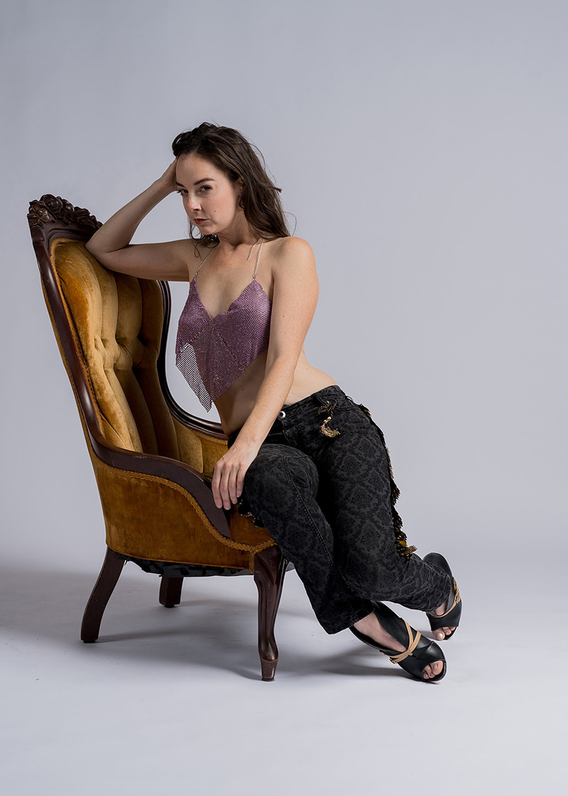 Young woman sitting sideways on a vintage mustard-yellow velvet high-back chair, wearing a lavender shimmering cropped top and black patterned pants with black sandals, against a plain light gray background.