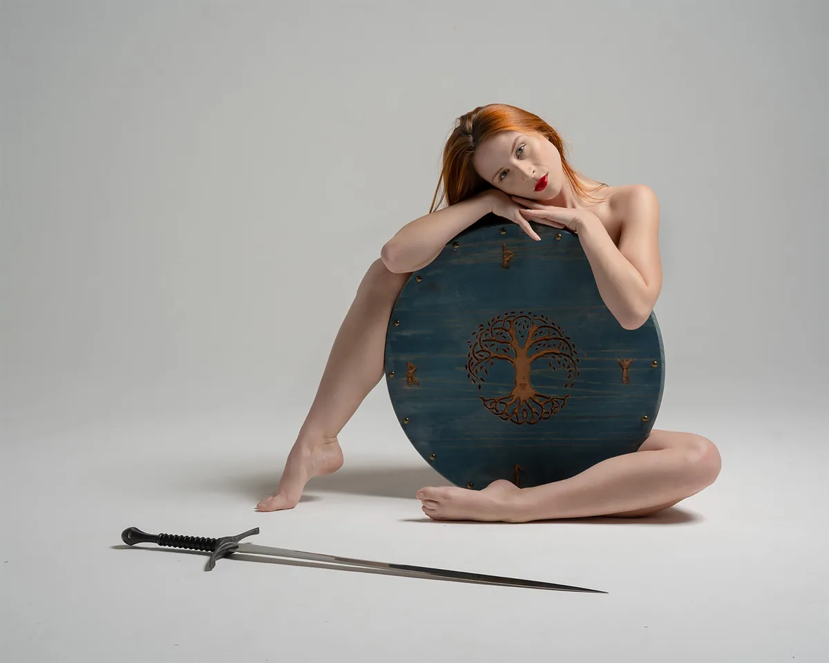 A woman with red hair and red lipstick sitting nude on the floor, leaning on a round blue shield with a carved tree design. A long sword with a black handle lies on the floor in front of her against a plain light background.