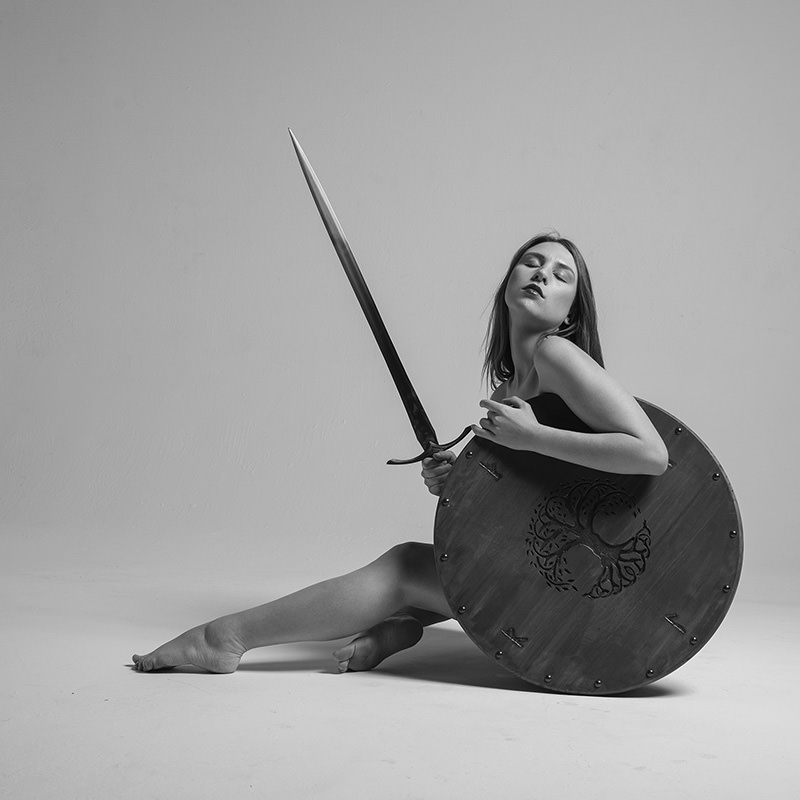 A woman sitting on the floor holding a round shield with a tree emblem and a long sword, eyes closed, in a black and white setting.