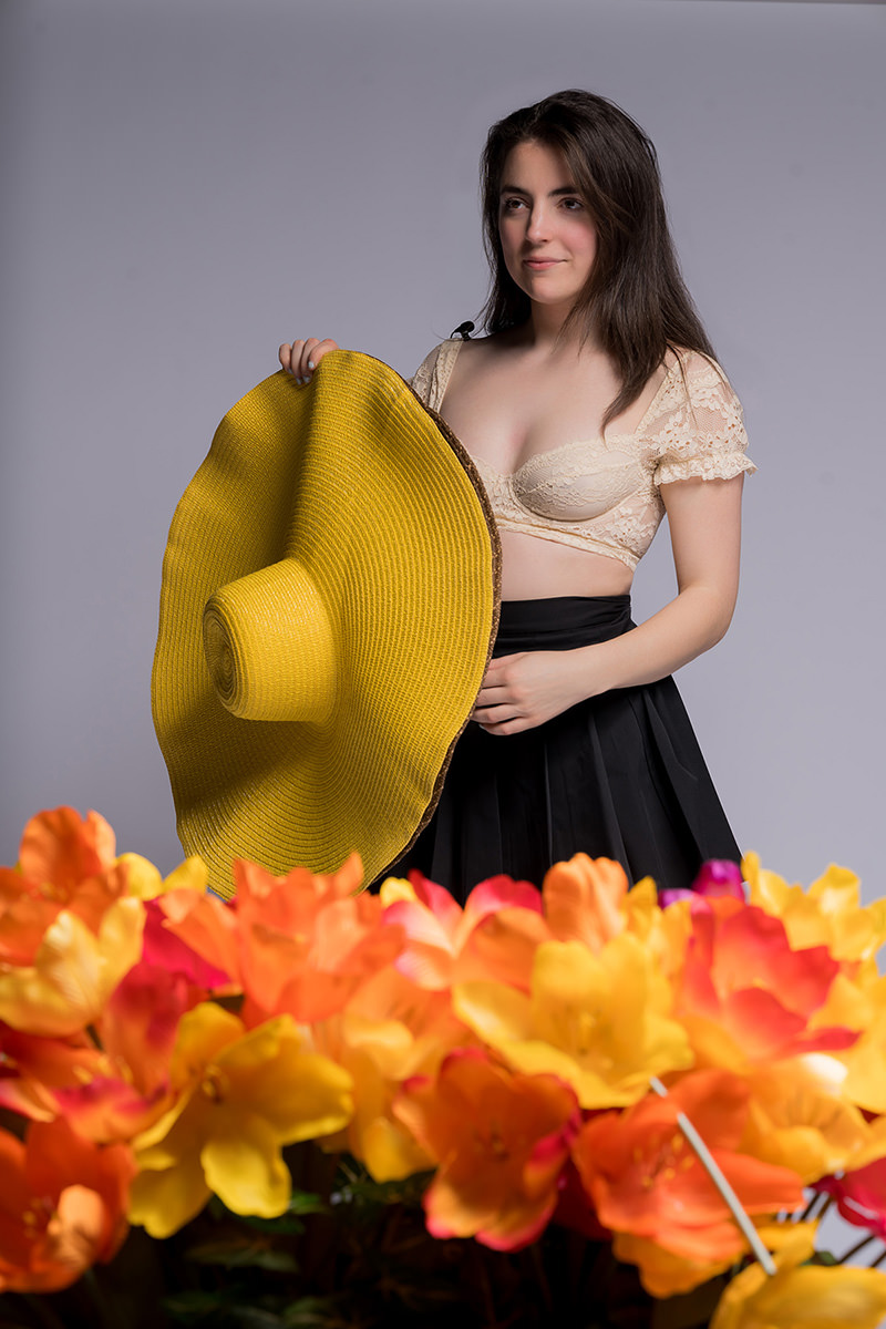 Young woman with long dark hair wearing a beige lace crop top and black skirt holding a large yellow wide-brimmed hat, standing behind a cluster of bright orange and yellow flowers against a plain gray background