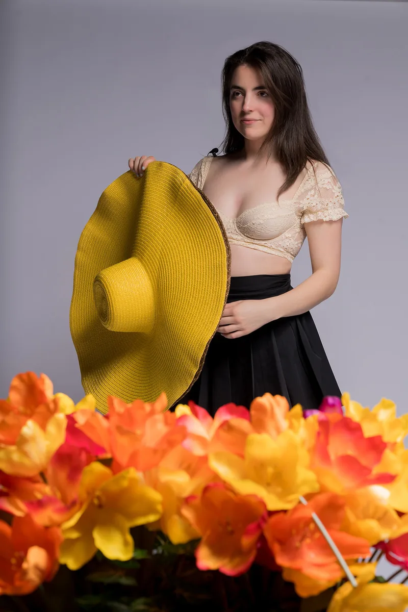 Young woman with long dark hair wearing a beige lace crop top and black skirt holding a large yellow wide-brimmed hat, standing behind a cluster of bright orange and yellow flowers against a plain gray background