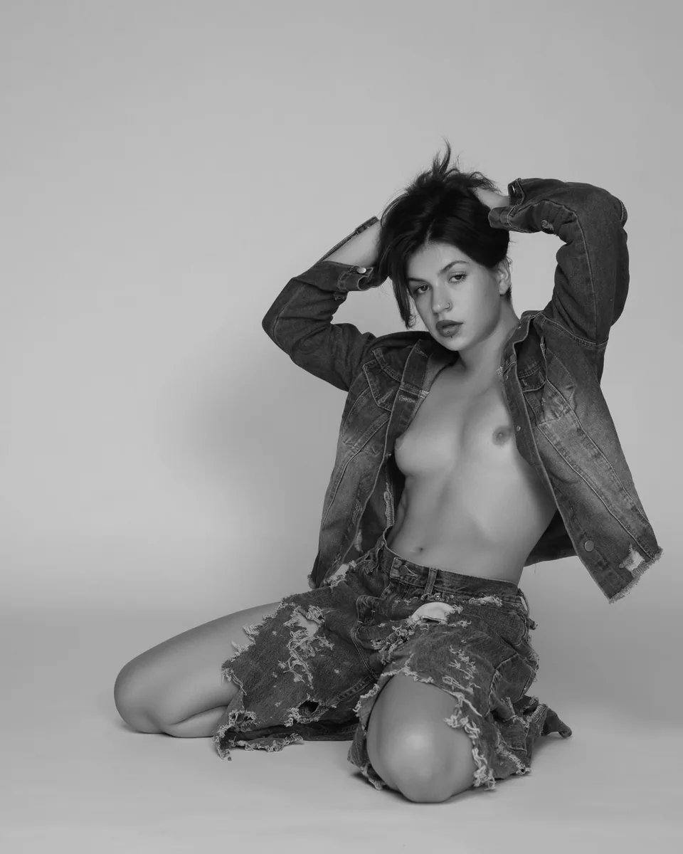 A young person kneels on a plain background wearing a distressed denim jacket and shorts, with arms raised holding hair, lit softly to emphasize calm expression and texture.