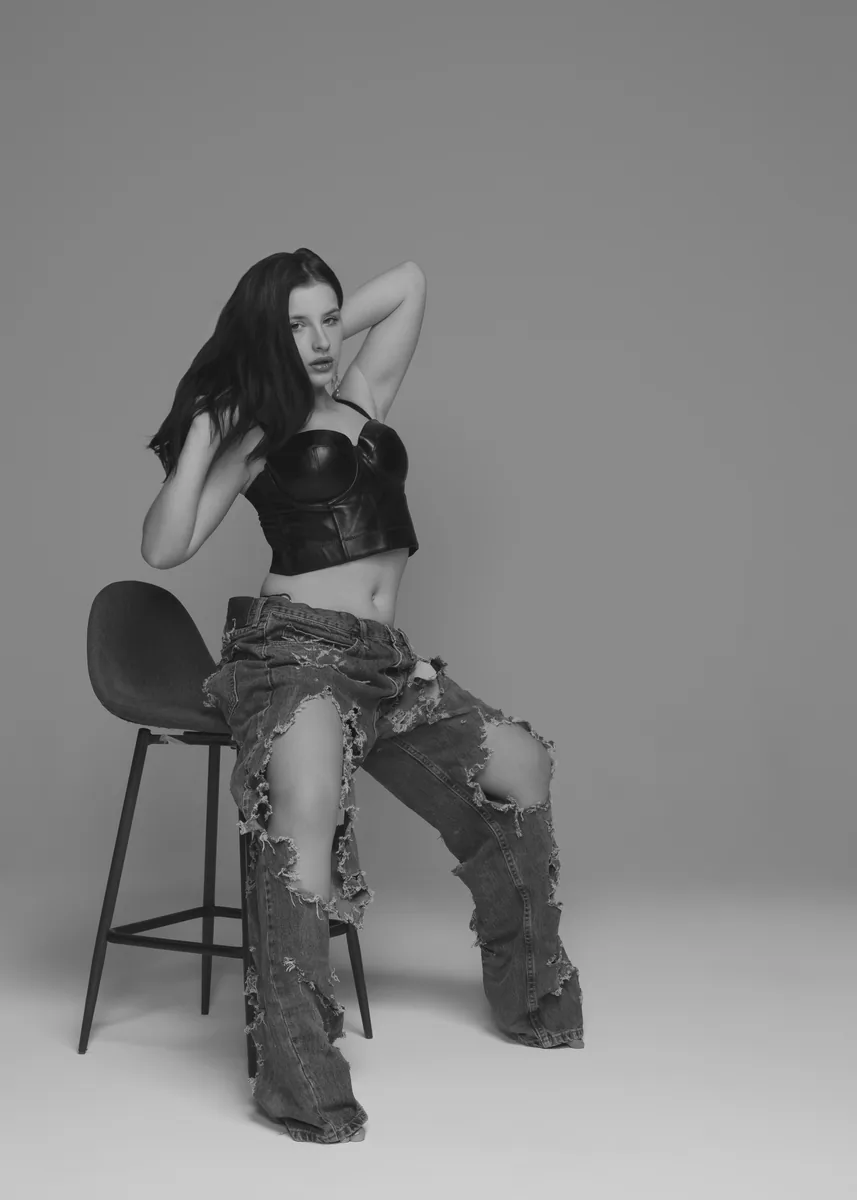 Black and white image of a seated woman with long dark hair wearing a cropped leather top and heavily torn wide-leg jeans, lit with soft even lighting against a plain background.