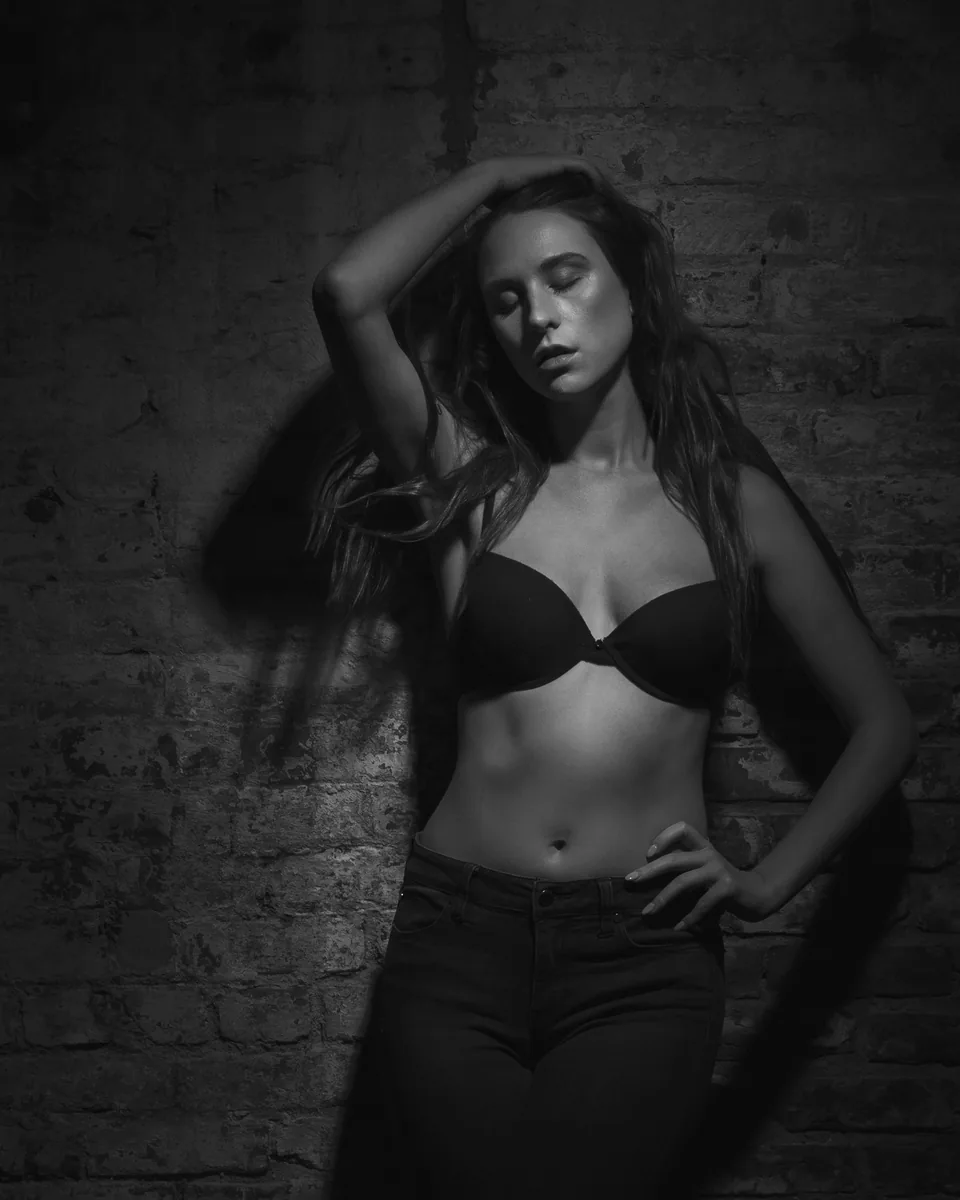 Black and white portrait of a woman standing against a textured brick wall, with one hand resting on her head and the other on her hip, illuminated by side lighting.