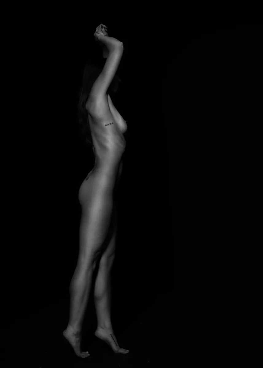 Black and white image of a standing woman in profile with arms raised above her head, softly lit against a dark background, revealing smooth skin and several small tattoos.