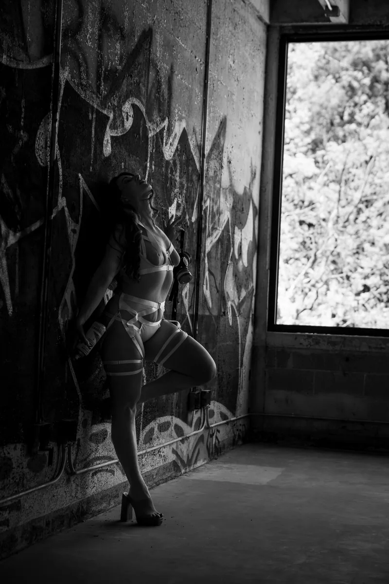 A woman in strappy lingerie and heels leans against a graffiti-covered wall, holding a spray can, with light coming from an open window illuminating her profile.