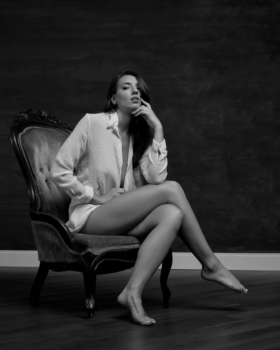 Black and white portrait of a woman seated on an ornate chair, wearing an unbuttoned textured shirt, with one leg crossed over the other and her hand resting near her chin.